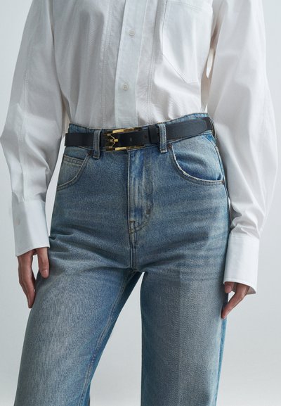 Μαύρη δερμάτινη ζώνη με χρυσό μεταλλικό κούμπωμα, φορεμένη πάνω από ανοιχτό μπλε τζιν παντελόνι, συνδυασμένη με λευκό πουκάμισο με κουμπιά.