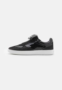 Svart sneaker med en kombinasjon av glatt patentlær og teksturert semsket skinn. Hvit gummisåle, svarte lisser, og logodetalj på siden.