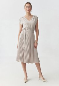 Abito beige plissettato con scollatura a V e maniche a cappuccio, caratterizzato da una vita legata e un tessuto liscio. Abbinato a décolletés a punta.