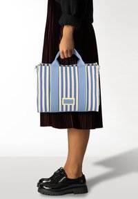 Gestreepte tote bag van canvas met lichtblauwe en gele strepen, voorzien van twee handvatten aan de bovenkant, een ritssluiting en een rechthoekig logo detail.