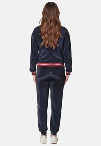 Marineblaue Velours-Jogginganzug mit einer cropped Jacke und schmal zulaufenden Hosen. Mit roten und weißen gestreiften Rippbündchen an den Ärmelabschlüssen und am Bund.