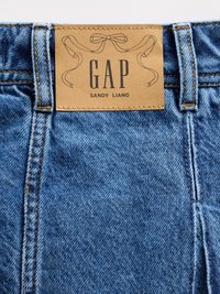 Jeans en denim bleu clair avec une étiquette marron portant les inscriptions "GAP" et "SANDY LIANG". Présente des coutures orange et des coutures visibles, avec une texture lisse.