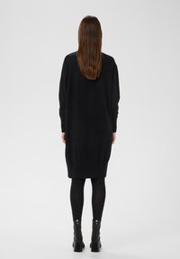 Schwarzes Strickkleid mit langen Ärmeln und abgerundetem Saum, ausgestattet mit einem gerippten Taillenbund. Kombiniert mit schwarzen knöchelhohen Stiefeln mit hohen Absätzen.