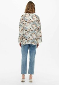 Blusa con stampa floreale color crema con fiori blu e marroni, vestibilità rilassata, maniche lunghe. Abbinata a jeans azzurri chiari e tacchi bianchi.