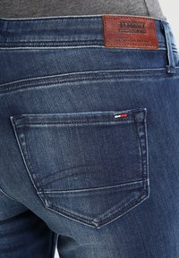 Vaqueros de mezclilla azul oscuro con un parche de cuero en la parte posterior etiquetado como "TOMMY JEANS". El diseño incluye bolsillos traseros con costuras sutiles.