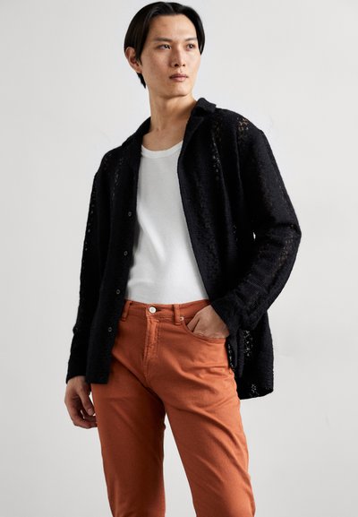 Svart stickad kofta med knappar, lagd över en vit skjorta. Matchad med orange byxor, med en framficka och synlig sömnad.