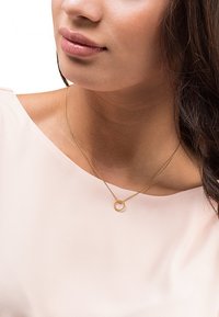 Collana d'oro con una catena delicata e un pendente circolare con un design testurizzato, indossata su un top chiaro.