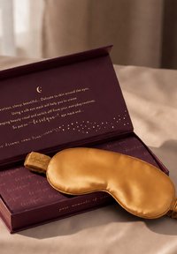 Seiden-Augenmaske in Gold, liegend auf einer offenen violetten Schachtel mit goldener Schrift, die zarte Hautpflege und Entspannungs-vorteile bewirbt.
