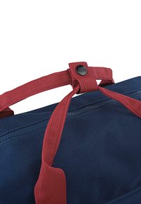 Bolso azul marino con un tejido texturizado, asas rojas y un botón de marca negro. La parte superior se cierra con una correa y un broche a presión.