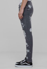 Vaqueros de mezclilla gris de corte slim con detalles de salpicaduras de pintura y acentos desgastados. Combinados con zapatillas negras de corte bajo con cordones blancos.