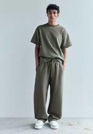 Tânăr purtând un tricou kaki larg și pantaloni largi asortați, cu adidași albi, stând în fața unui perete simplu, deschis la culoare.