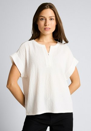 Kobieta o długich brązowych włosach, ubrana w biały top o fakturze z krótkim rękawem i czarne spodnie, stojąca ze złączonymi rękami za plecami na szarym tle.