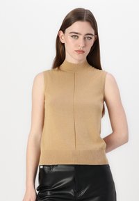 Beige top bez rukávů s rolákem a přiléhavým střihem, se žebrovaným límcem. Předvedený s černými kalhotami z umělé kůže.