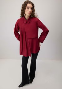 Punane nööpidega särk, millel on ühel küljel voltidega disain. Pika varrukaga ja kraega. Kombineeritud mustade lehvikpükste ja teravnurksete kingadega.