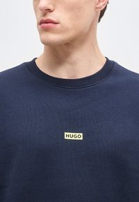 Νέος άνδρας που φοράει μπλε σκούρο φούτερ με στρογγυλή λαιμόκοψη, με ένα μικρό ορθογώνιο λογότυπο "HUGO" στο κέντρο του στήθους.