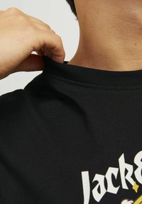 Juodas medvilnės marškinėliai su apvaliu kaklu. Ranka šiek tiek tempia apykaklę, pabrėždama siuvimo detalę ir tinkamumą. Matomas ryškus baltas ir geltonas tekstas.