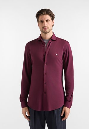 Uomo che indossa una camicia bordeaux a maniche lunghe con un piccolo logo bianco di un cane sul petto e pantaloni scuri, in piedi davanti a uno sfondo bianco.