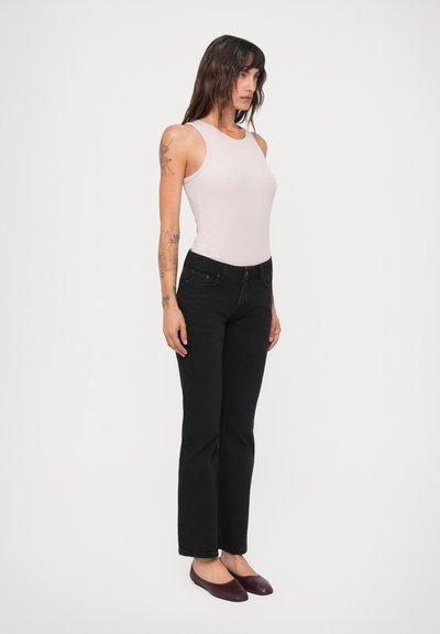 Kvinde med langt mørkt hår, tatoveringer på armene, iført en lys pink ærmeløs top, sorte jeans og mørkerøde flade sko, stående i profil.