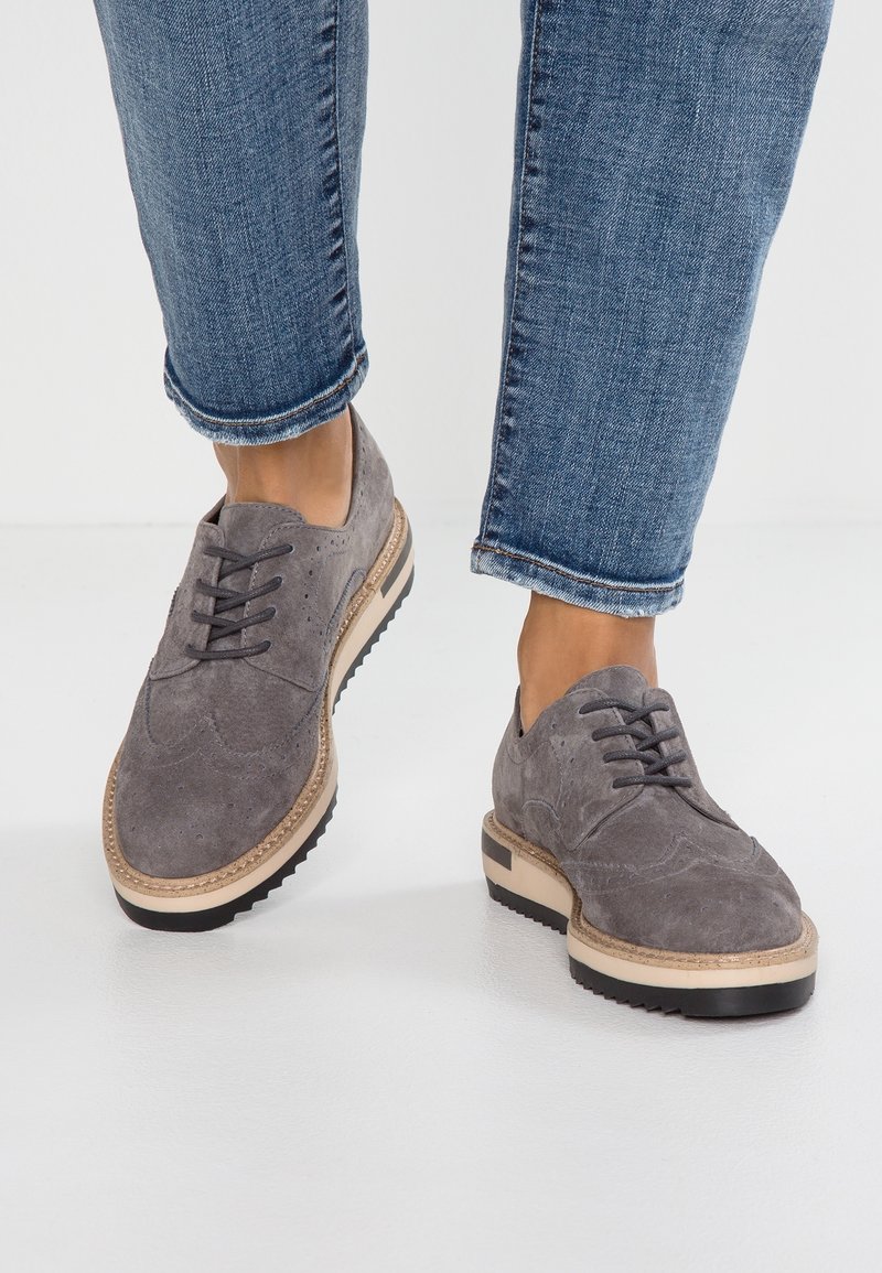 Personne portant un jean bleu retroussé et des chaussures brogues en daim gris avec des semelles épaisses beige et noire, debout sur un sol blanc.