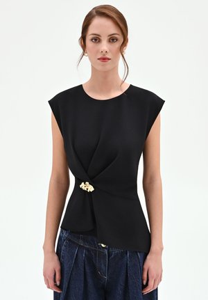 Femme portant un haut noir sans manches avec détail de fermoir doré à la taille et un pantalon ample plissé bleu foncé sur fond blanc.