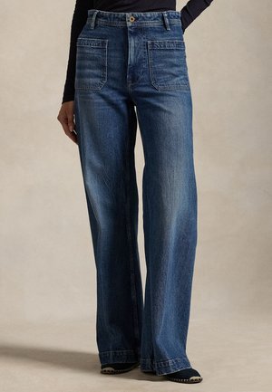 Modèle portant un jean bleu foncé à jambes larges, avec deux grandes poches plaquées à l’avant, et un haut noir sur un fond neutre.