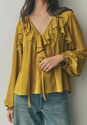 Kvinde iført sennepsgul bluse med flæset V-hals og lange, puffede ærmer kombineret med blå jeans, hånd hvilende nær taljen.