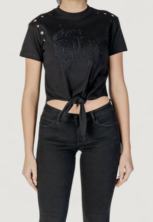 Kvinde iført sort binde-crop top med præget logo og øjendetaljer kombineret med sorte højtaljede jeans mod en ensfarvet baggrund.
