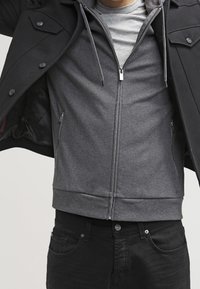 Sweat-shirt gris à fermeture éclair avec une capuche, doté de poches latérales, d'une texture lisse et d'une coupe ajustée, porté sous une veste noire.