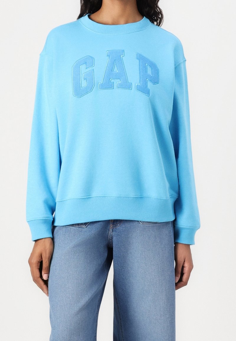 Personne portant un sweat GAP bleu clair avec des lettres en relief du logo et un jean bleu moyen, sur un fond blanc uni.