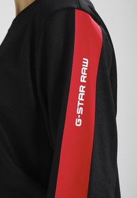 Sudadera negra con una llamativa franja roja a lo largo de la manga izquierda con "G-STAR RAW" impreso en blanco. Textura de tela suave, diseño casual.