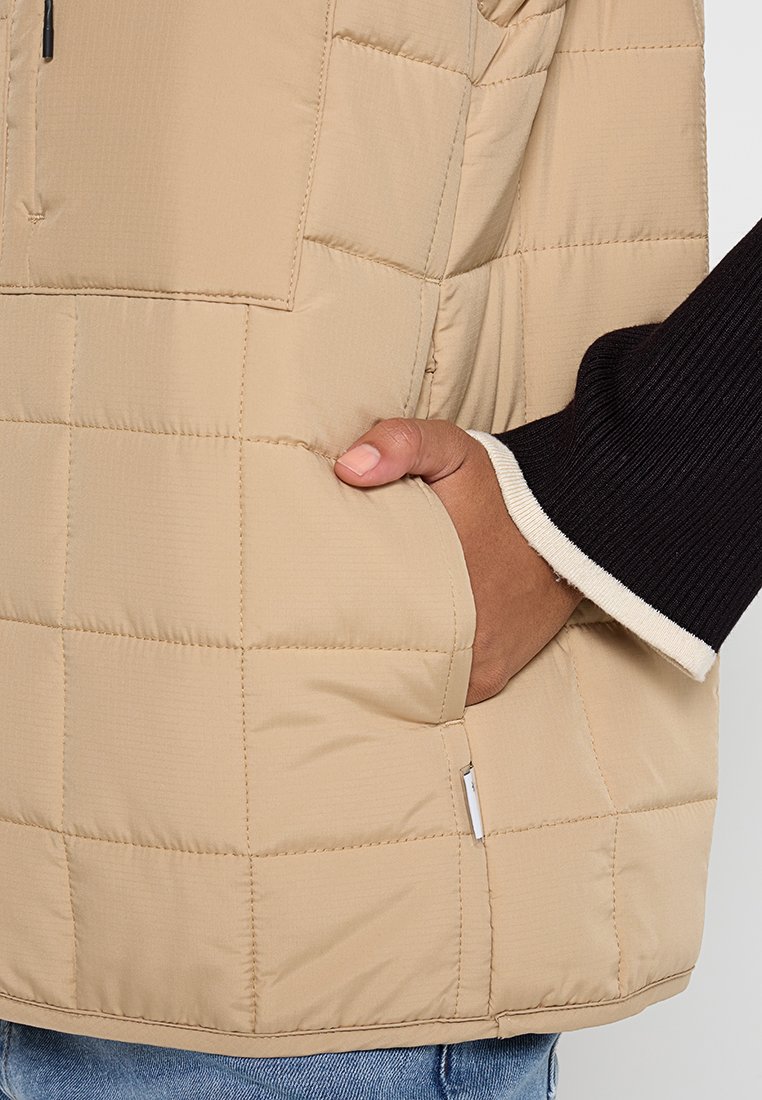 Beige gesteppte Jacke mit einer Vorderseite Tasche, glatter Textur, halben Reißverschluss und kontrastierendem schwarzen Rippbündchen mit weißen Akzenten.