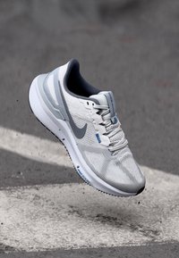 Grå og hvit joggesko med mesh overdel, teksturert design, Nike-logo og blå detaljer. Har en dempet såle og snøring.