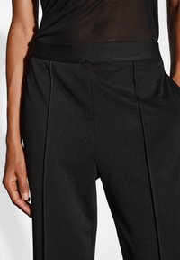 Pantalons noirs en tissu lisse et texturé, avec une large taille élastique et des plis à l'avant. Le design est minimaliste et ajusté.