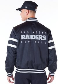 Chaqueta bomber negra de satén con rayas blancas, que presenta "LAS VEGAS RAIDERS FOOTBALL" en letras gris oscuro en la parte de atrás. Cuello y puños de canalé.