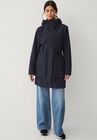 Marineblauwe waterdichte jas met een capuchon, voorzien van een ritssluiting, twee voorkzakken en een verstelbare trekkoord in de taille. Gecombineerd met wijde jeans.