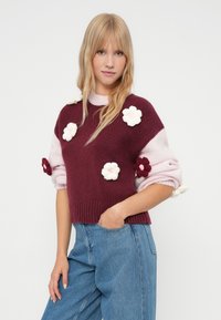 Pulover tricotat cu un corp mov, mâneci roz și aplicații cu flori. Are un tiv elastic și un material texturat. Asociat cu blugi din denim albastru.