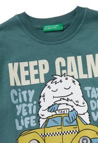 Tyrkysové bavlněné tričko s karikaturou yetiho s modrými drápy, který drží žluté taxi, a textem "KEEP CALM CITY YETI LIFE" v výrazných barvách.