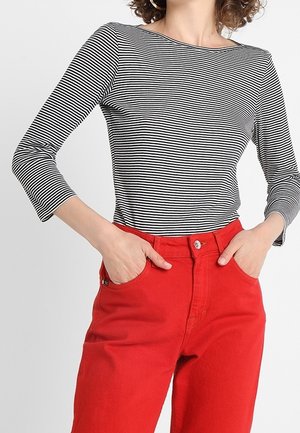 Vrouw draagt een zwart-wit horizontaal gestreept shirt met lange mouwen, ingestopt in felrode high-waisted broek, met handen in de zakken.