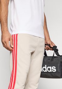 Vyras baltame marškinėlyje ir smėlio spalvos kelnėse su trimis raudonomis šoninėmis juostelėmis laiko mustangų maišelį „Adidas“ juo laikydamas už rankenos.