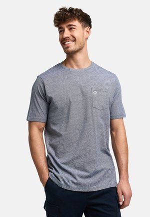 Smilende mand iført en grå kortærmet t-shirt med rund hals og en lille brystlomme samt mørke bukser, stående med hænderne i lommerne.
