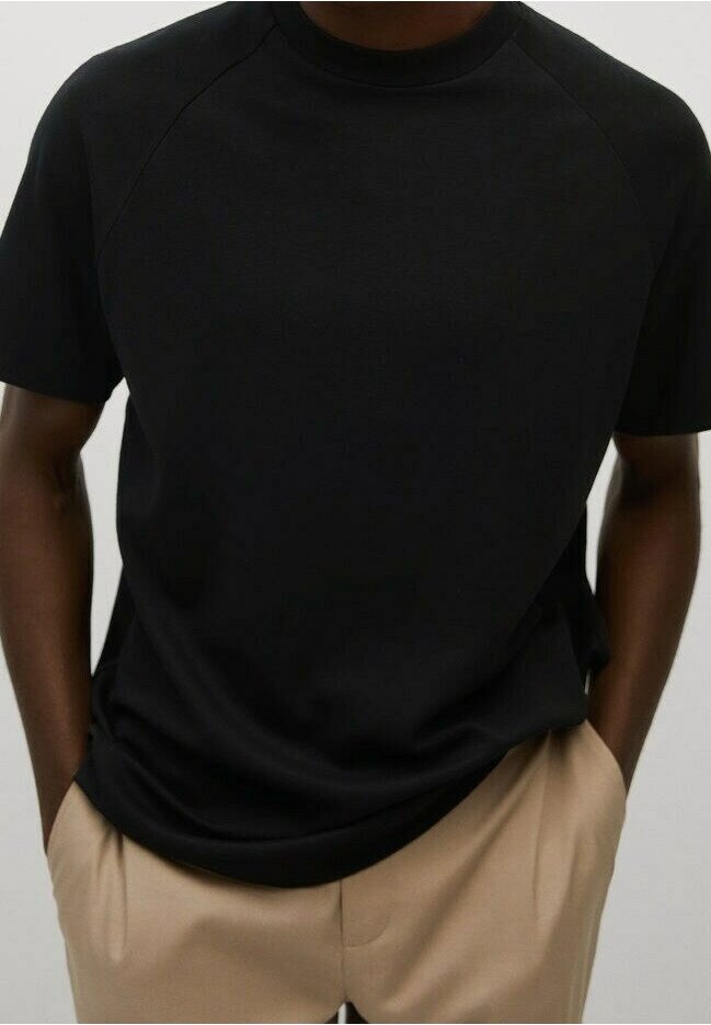 T-shirt en coton noir avec col rond et manches raglan courtes. Texture lisse, coupe légèrement ample, associé à un pantalon beige.