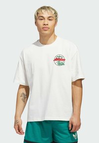 Bijela pamučna majica kratkih rukava sa šarenim grafičkim logotipom na lijevoj strani prsnog koša. Nosena je uz tirkizne atletske kratke hlače.