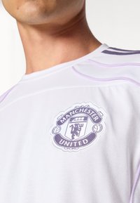 Hvid atletisk skjorte med et lilla Manchester United-logo broderet på venstre bryst. Afsluttet med diskrete lilla syninger.