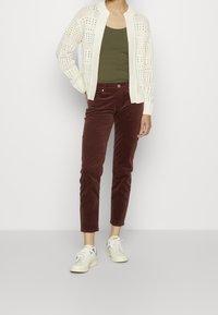 Osoba oblečená do bílé otevřené pletené kardiganu, olivově zeleného tílka, hnědých kordových kalhot a bílých tenisek s modrými akcenty, stojící na jednobarevném pozadí.