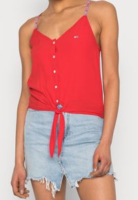 Top rojo sin mangas con detalle de lazo en la parte delantera, cierre de botones y tirantes en contraste. Combinado con una falda de mezclilla desgastada en azul claro.