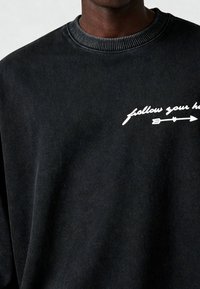 Zwart crewneck sweatshirt met witte tekst "follow your he" en een naar rechts wijzende pijl op de borst.