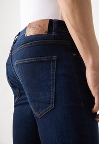 Mörkblå denimjeans med en figursydd design, som har kopparstickningar, två bakfickor och en tanfärgad läderlapp med logotyp på midjebandet.
