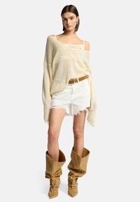 Pull en tricot beige clair surdimensionné, shorts blancs effilochés et bottes montantes en tan à motifs avec bouts pointus, dotées de détails cloutés.