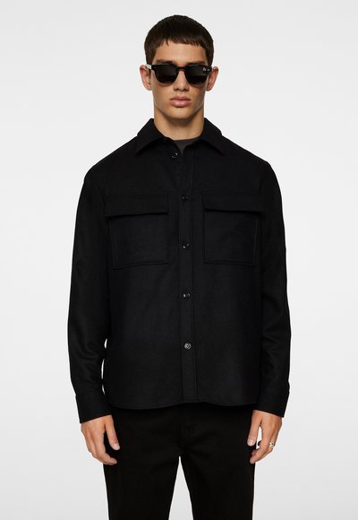 Homme debout portant une chemise noire boutonnée avec deux poches poitrine, des lunettes de soleil noires et un pantalon noir sur un fond clair uni.