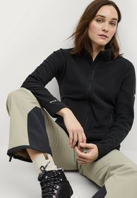 Zwarte fleecejack met hoge kraag en ritssluiting aan de voorkant. Bruine broek met zwarte accenten. Zwarte wandelschoenen en gemusterde sokken zijn zichtbaar.
